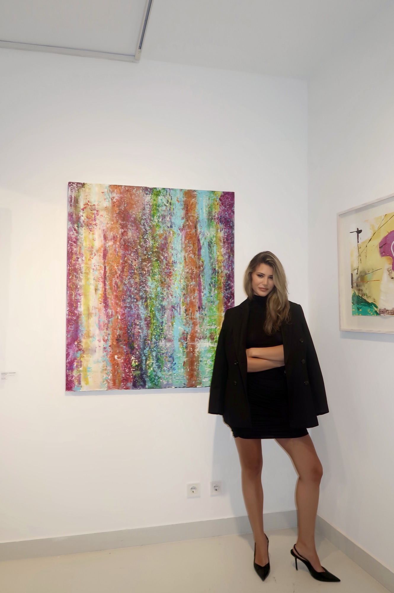 Sydal standing beside her large abstract painting on the white wall of The Holly Art Gallery.